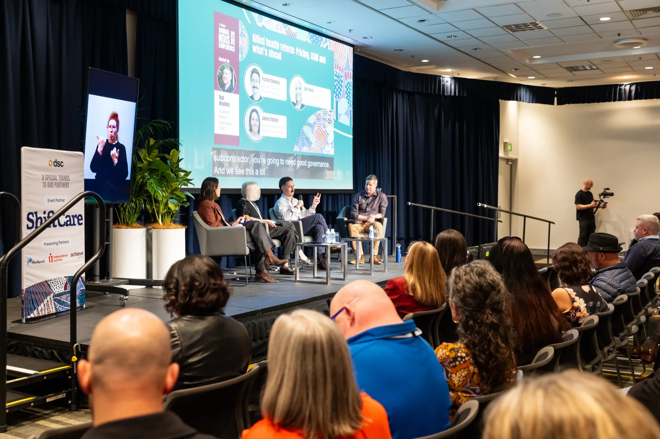 A DSC Annual NDIS Conference session seen from the back of the room, with four panellists on a lit stage, an Auslan interpreter on a screen beside them and a seated audience in the foreground.