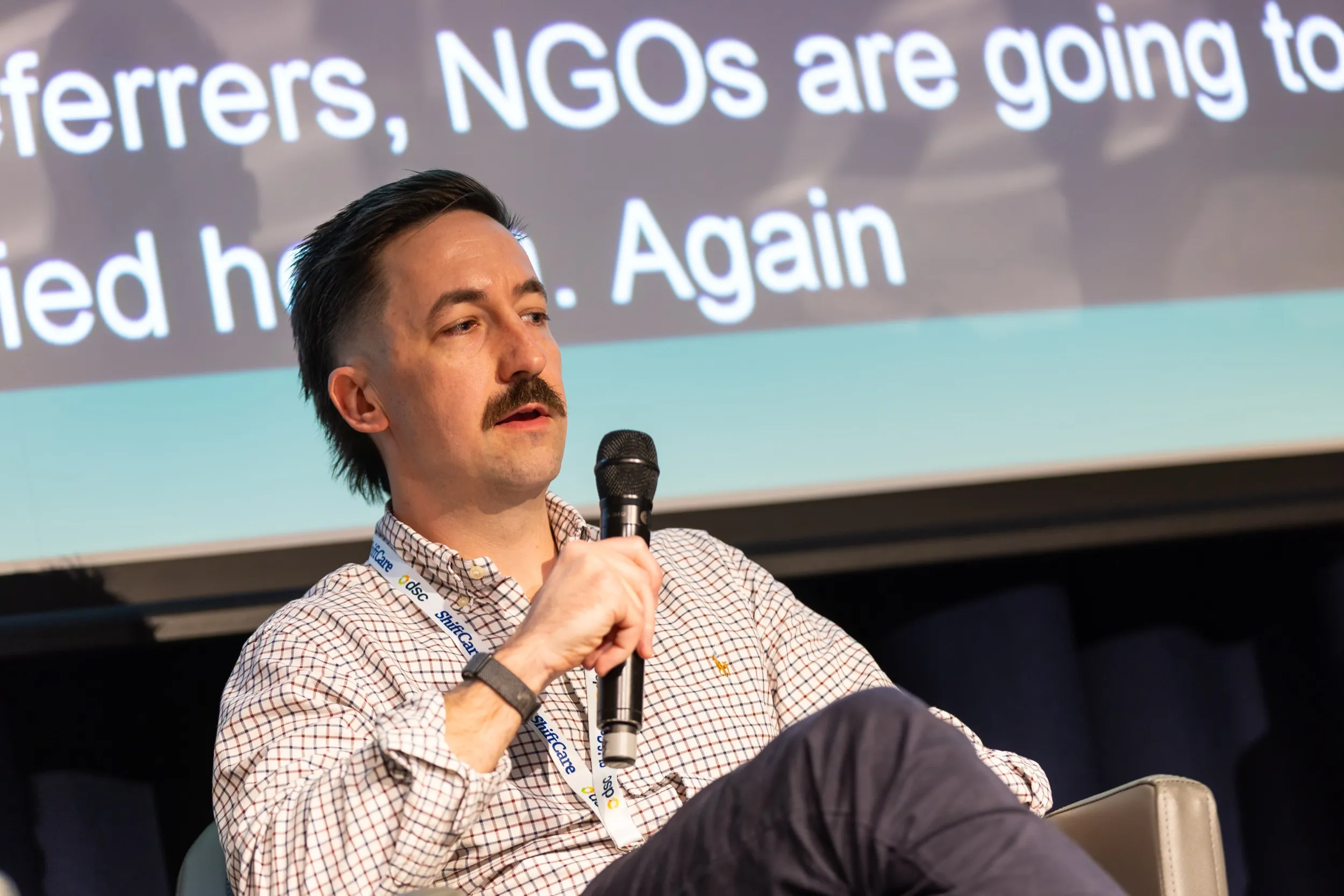Trystan Conway seated on stage at the DSC Annual NDIS Conference, speaking into a handheld microphone, with the session's live captions projected on the screen behind him.