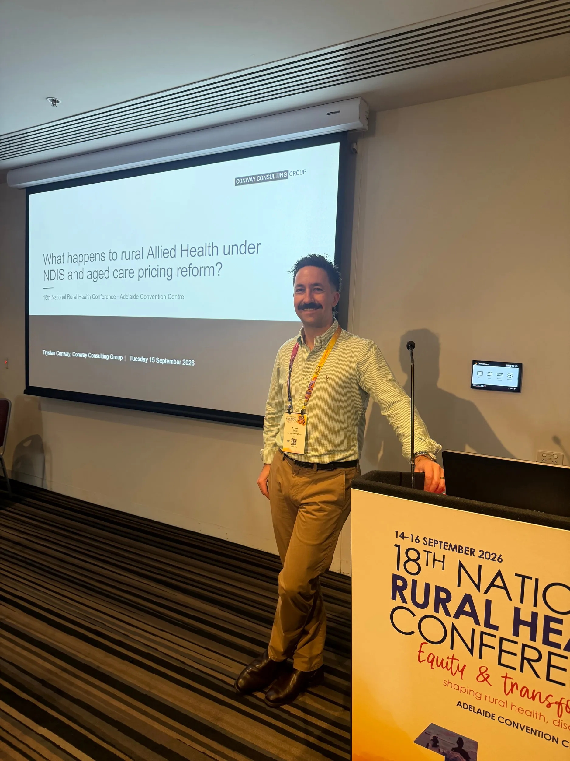 Trystan Conway standing beside the lectern at the 18th National Rural Health Conference, with his title slide on the screen behind him.